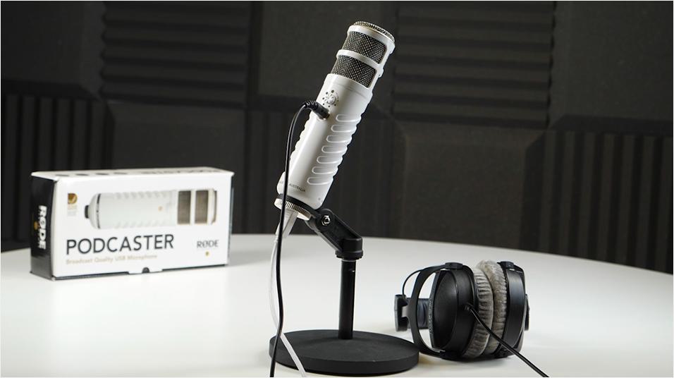 The RODE podcaster, on a mic table stand and next to a pair of headphones. The Podcaster box is at the back of the table in the background.