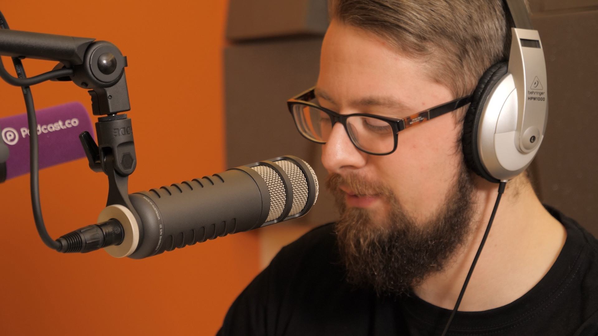 A man wearing headphones and talking into a black Rode Procaster microphone which is held by a boom arm.