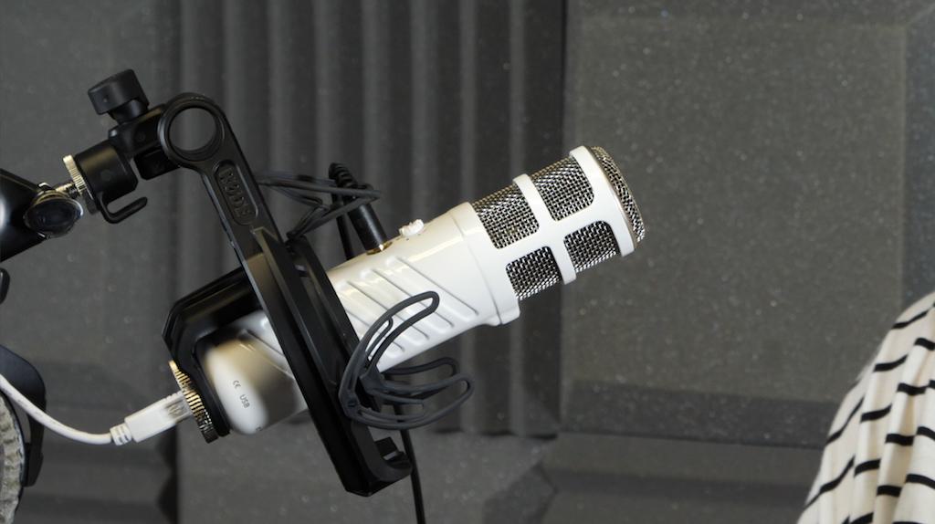 Image shows the white Rode Podcaster microphone hanging from a microphone stand in a radio studio.