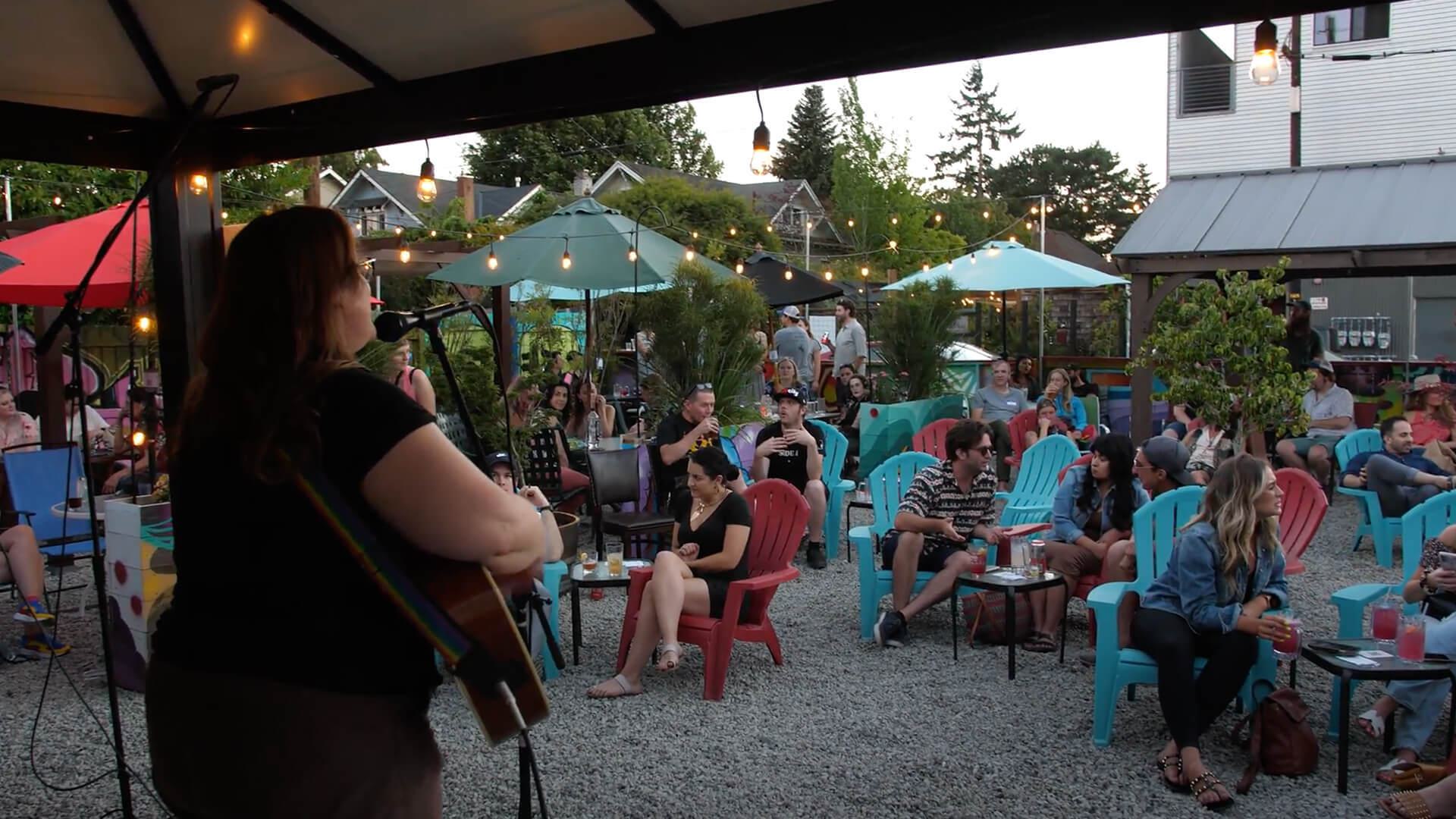 Image shows a woman playing guitar to a crowd outside at the Shady Pines Radio open mic