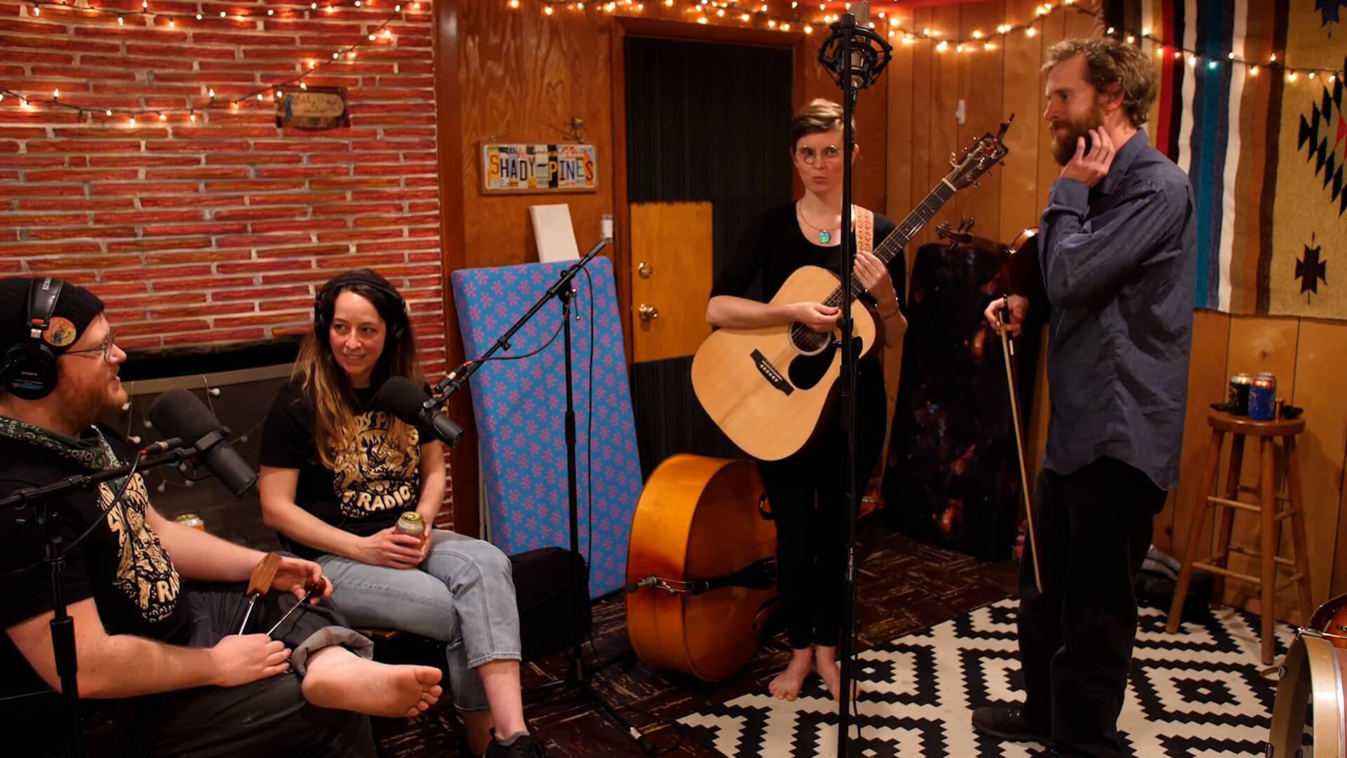Image shows the Shady Pines Radio band in session. Two instrumentalists are standing up and two singers are sat on a sofa.