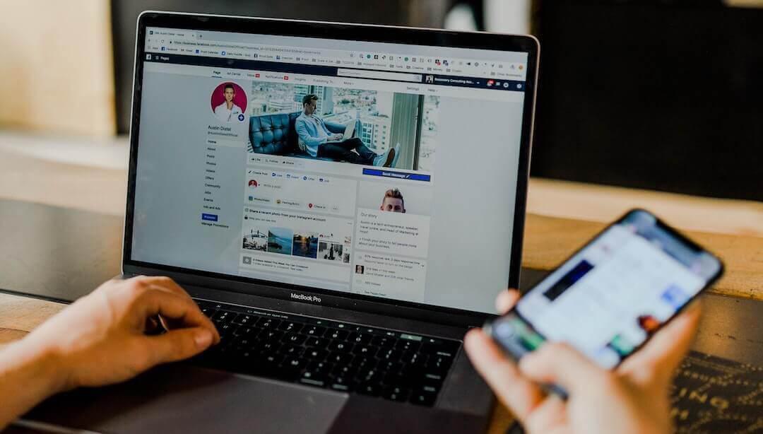 This is a photo of someone using social media on a Macbook in one hand, and their other hand is holding up a phone on social media. The background is neutral.