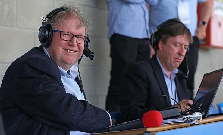 Two sports commentators in a stadium, both on laptops and wearing headphones.