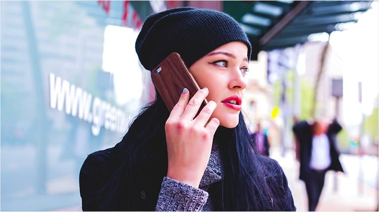 This is an image of a white woman with no hair visible under her hat wearing red lipstick. She has a black beanie hat on and a black coat, and is talking into an iPhone.