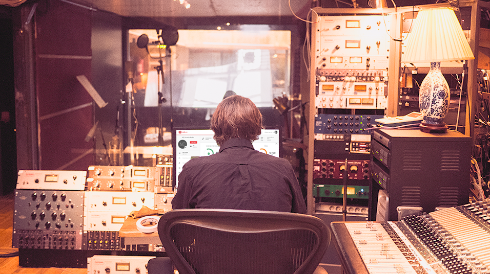 This is an image of a busy radio studio. There is a lamp on the left, warm lighting, and radio mixers and interfaces in silver on the lift. In the centre is a man from behind with greyish hair looking at a computer monitor.