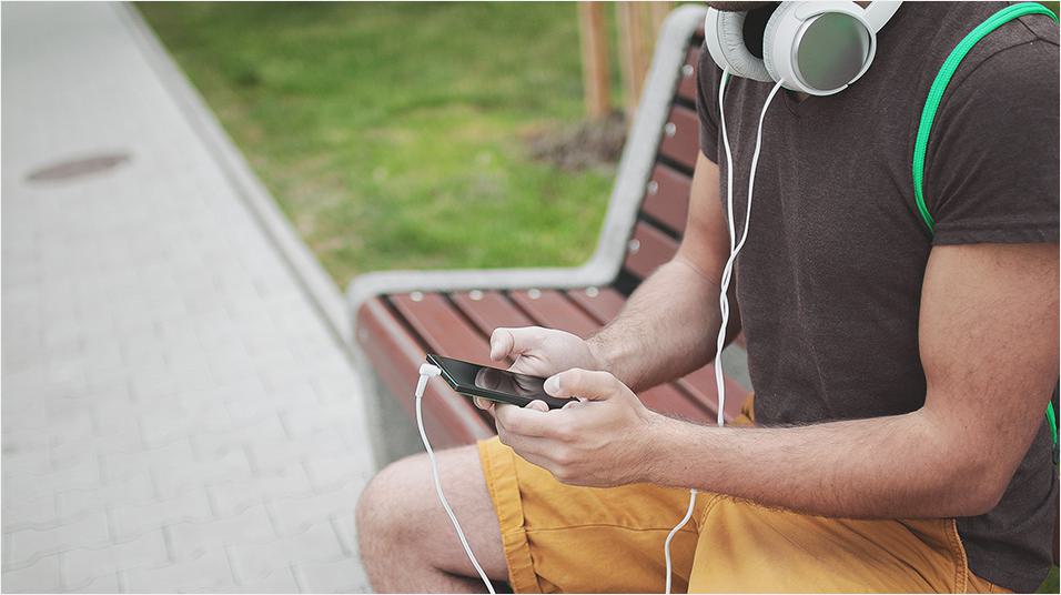 Image shows a man on a bench wearing headphones which are plugged into his smartphone.